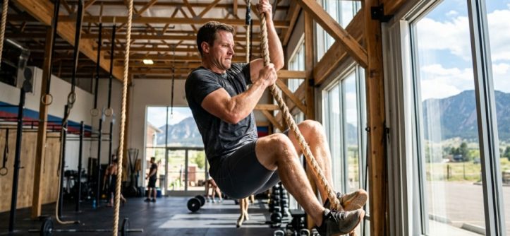 Athlete climbing a heavy-duty gymnastics rope inside a sunlit Colorado gym.